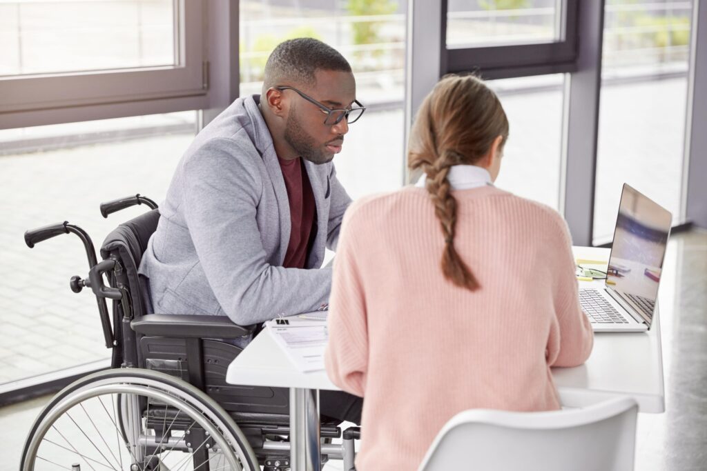 A person in a wheelchair and another person sitting at a desk with a laptop, engaged in what appears to be a consultation or meeting, resized