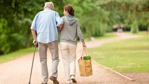 An older individual using a cane and accompanied by another person carrying groceries walk together on a serene, tree-lined path, resized